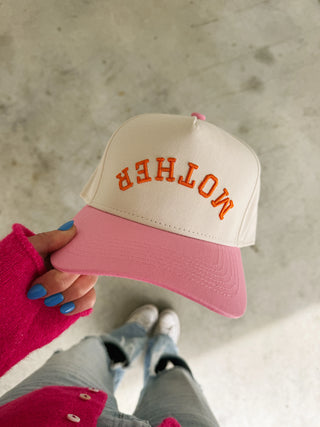 Pink and beige cap with 'MOTHER' text held by a person on a concrete floor.