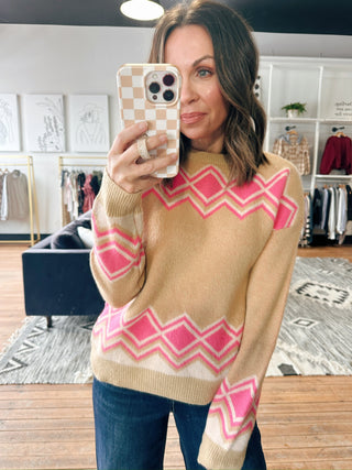 Woman taking a mirror selfie wearing a beige sweater with pink geometric patterns in a clothing store.