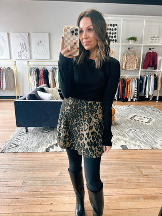 Woman taking a mirror selfie wearing a black top and leopard print skirt in a clothing store.