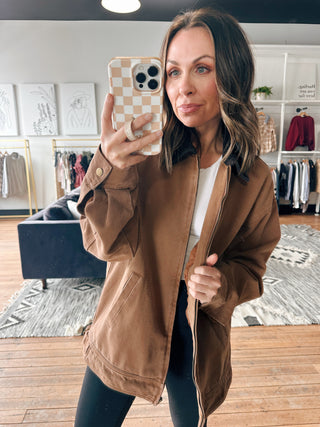 Woman taking a selfie in a brown jacket in a room with clothing racks.
