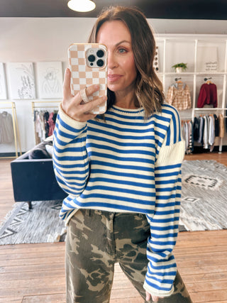 Woman taking a mirror selfie wearing a blue and white striped sweater and camouflage pants in a store.