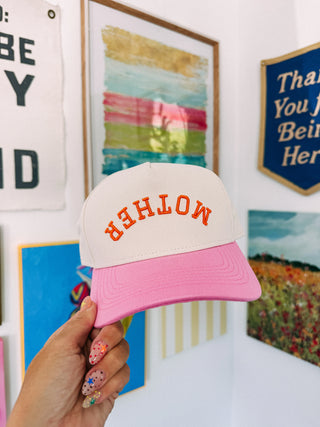 Pink and white cap with 'MOTHER' text held by a hand in front of wall art.